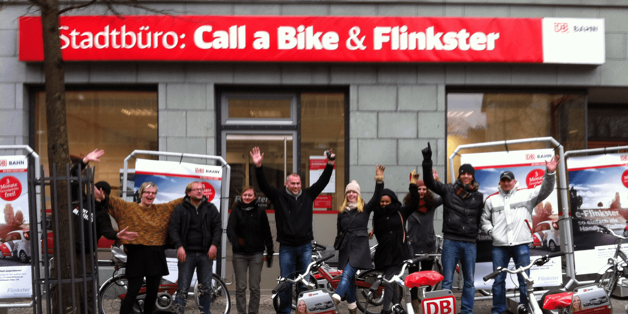 Cheerful group of people posing in front of a city office with the sign "Call a Bike & Flinkster", cheering and standing next to bicycles and advertising material for bicycle and car sharing services.