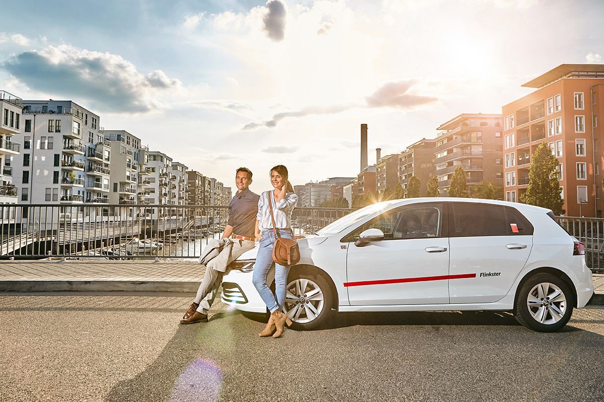 Ein Mann und eine Frau stehen entspannt an einem weißen Flinkster-Auto auf einer Brücke in einer modernen Stadt. Im Hintergrund sind Wohngebäude und Wasser zu sehen.