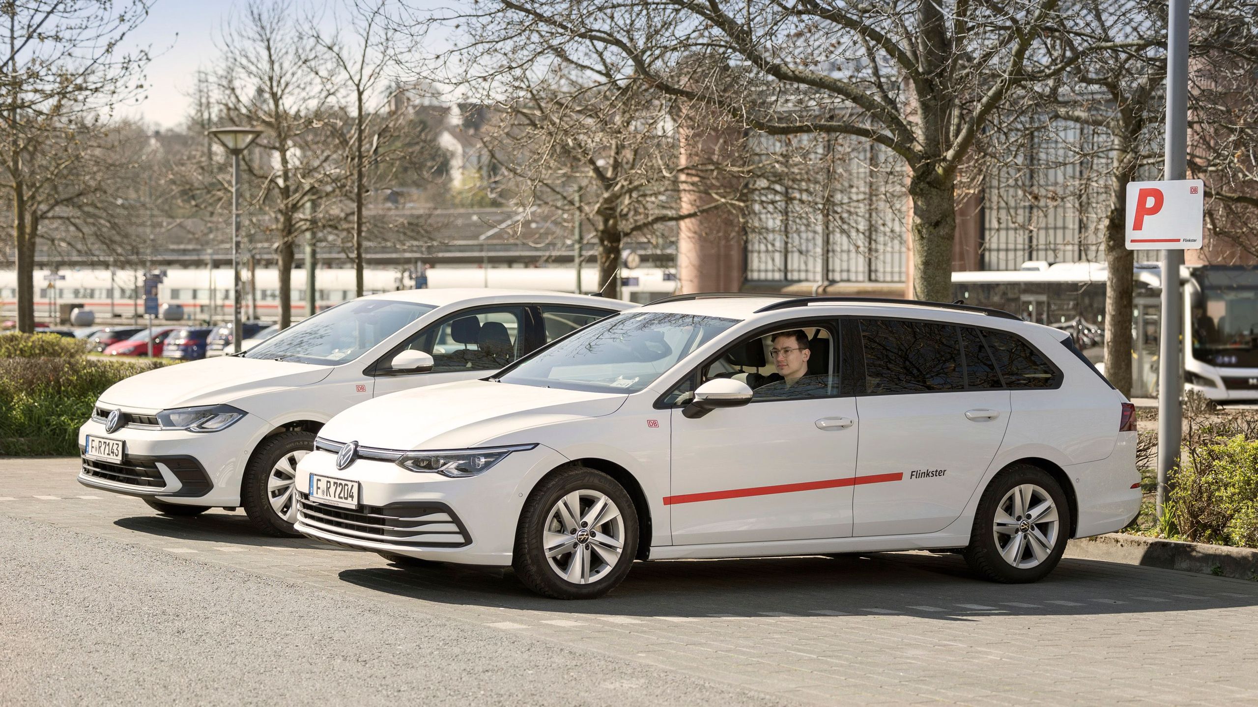 Das Bild zeigt zwei weiße Flinkster-Fahrzeuge auf einem Parkplatz, eines mit einem Fahrer. Im Hintergrund sind Bäume, geparkte Autos und ein Bahnhofsgebäude zu sehen. Ein Schild weist auf den reservierten Flinkster-Parkplatz hin.