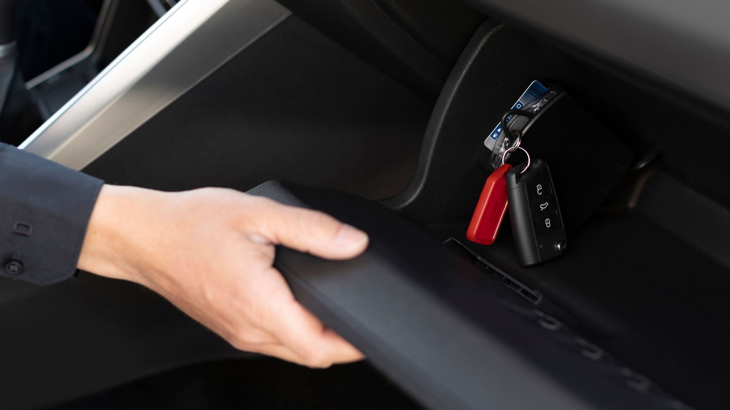 Innerraum von einem Auto mit von einer Hand geöffneter Fußraumklappe auf Beifahrersitz. Im Inneraum des Fachs ist der Fahrzeugschlüssel an einem Carsharing-Modul eingesteckt.