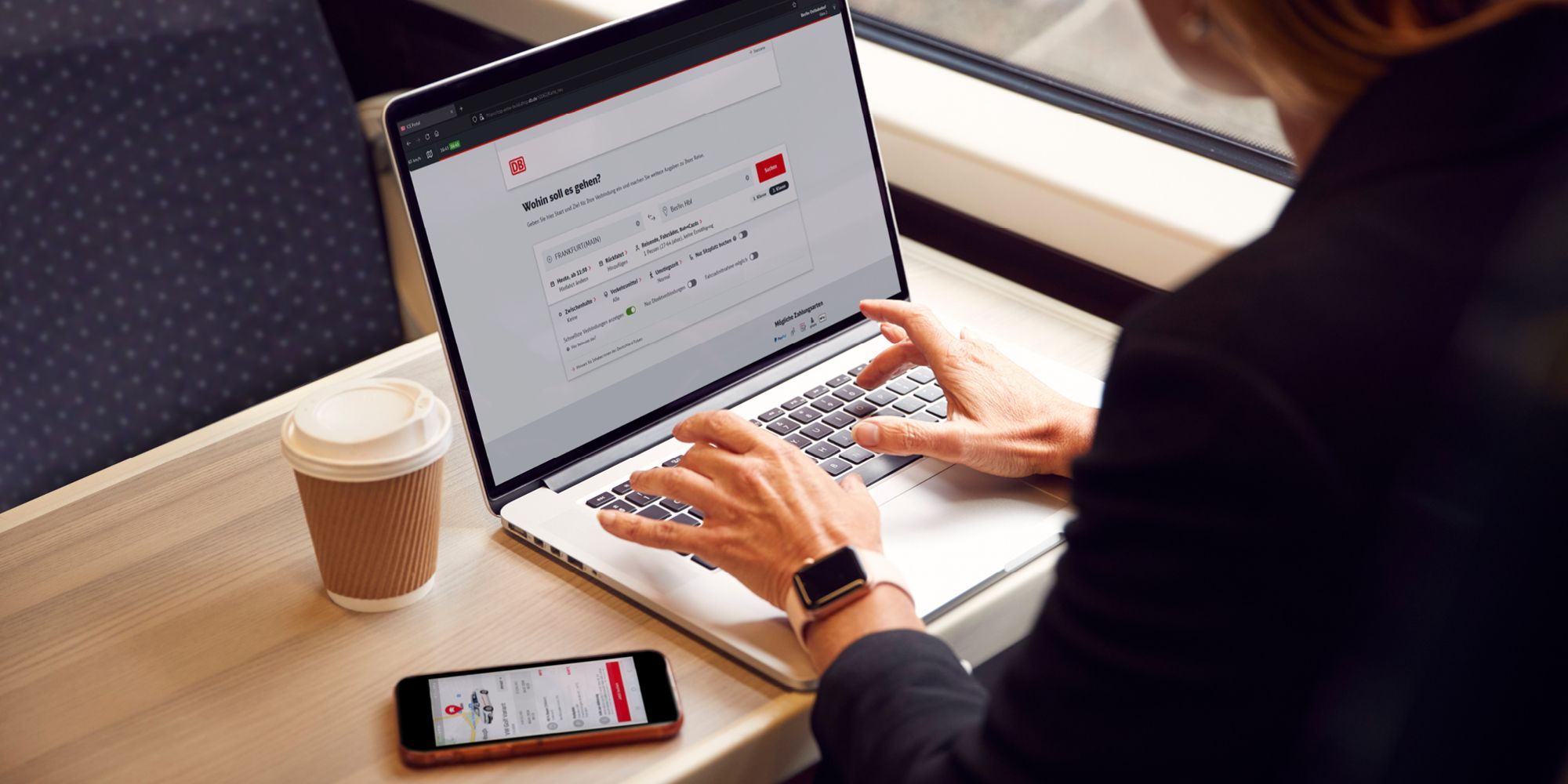 Eine Frau sitzt im Zug und arbeitet an einem Laptop, auf dem der DB Navigator geöffnet ist. Neben ihr steht ein Kaffeebecher und ein Smartphone, auf dem die Flinkster-App geöffnet ist.